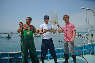 漁船の上で、3人の男性がそれぞれ、貝、魚、網を持って並んでいる写真