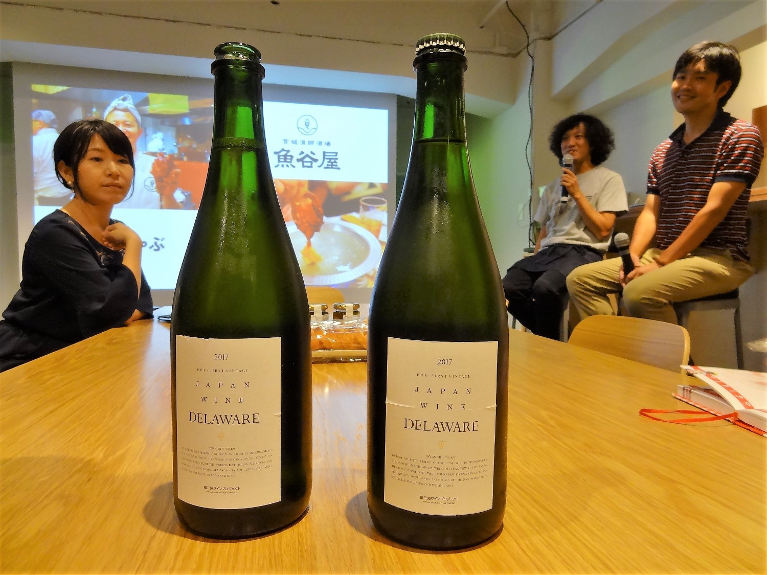 壁に映像が映し出され、横のカウンターの席にマイクを持って話をしている長谷川さん、正司さんが座る中、女性が座ったテーブルの上に2本置かれた「DELAWARE」と書かれたよく冷えた緑色のワインボトルにフォーカスを当てて撮影した写真