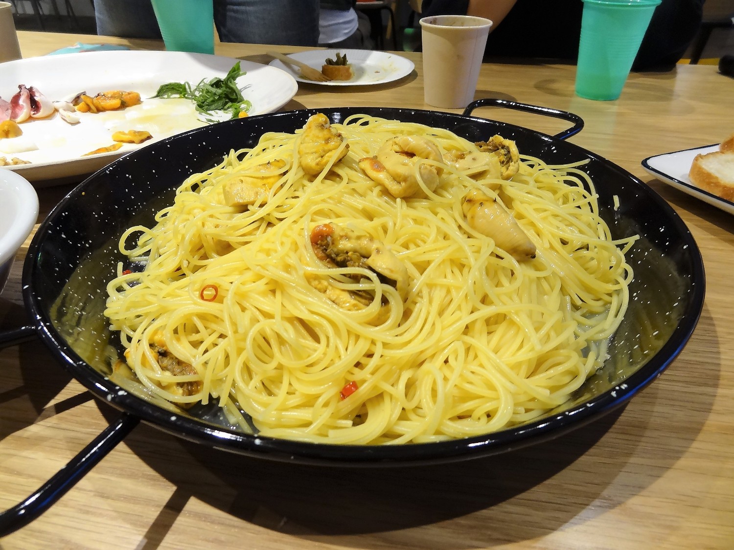 奥には食べかけの料理と緑のコップが見えるテーブルの上に、ホヤと赤唐辛子が入った、黄色い細めのパスタが黒い平たい鍋にたっぷりと盛られて置かれている写真
