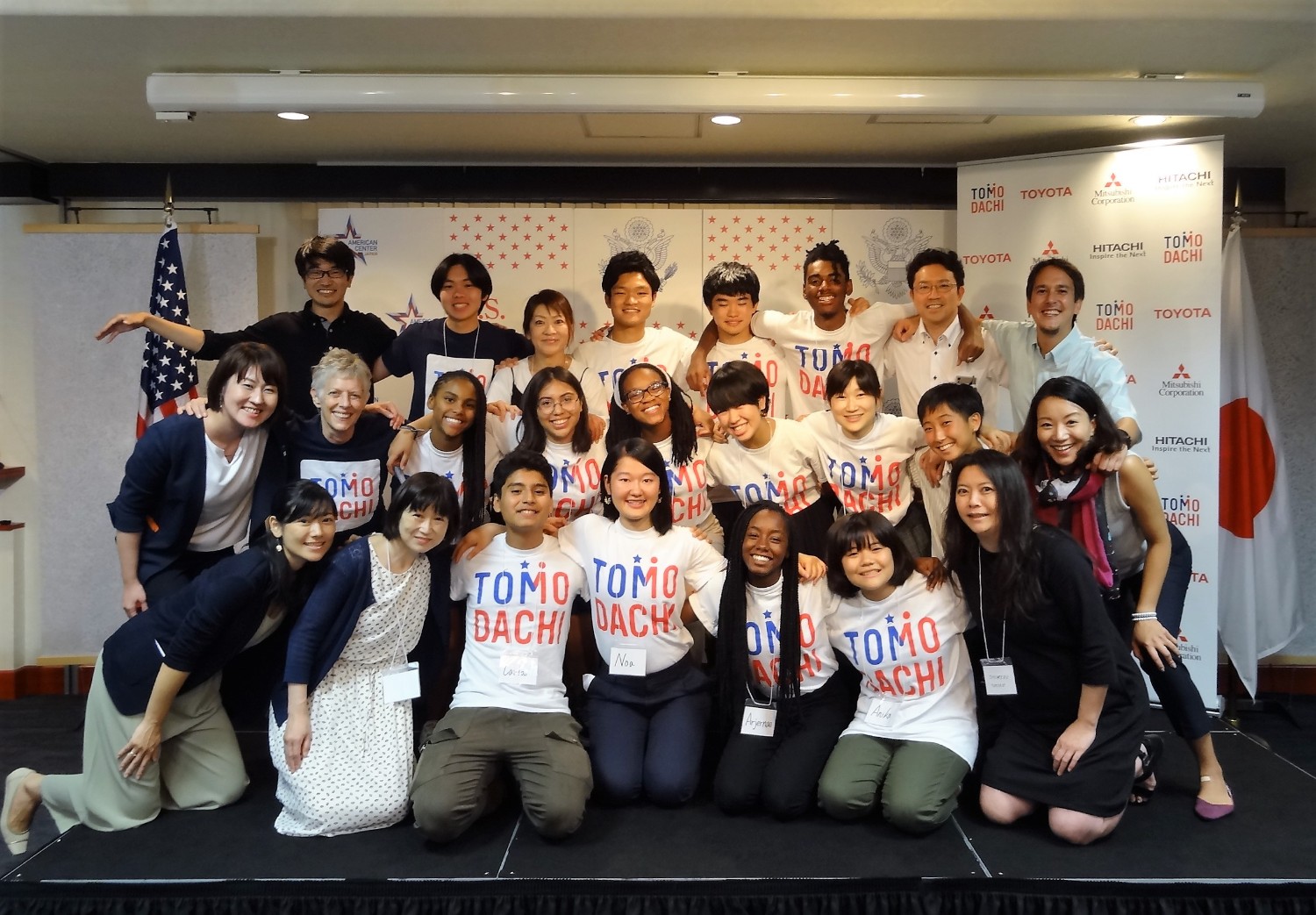 アメリカと日本の国旗の前で「TOMODACHI」Tシャツを着た日米の高校生たちが肩を組んで笑顔でポーズを取っている写真