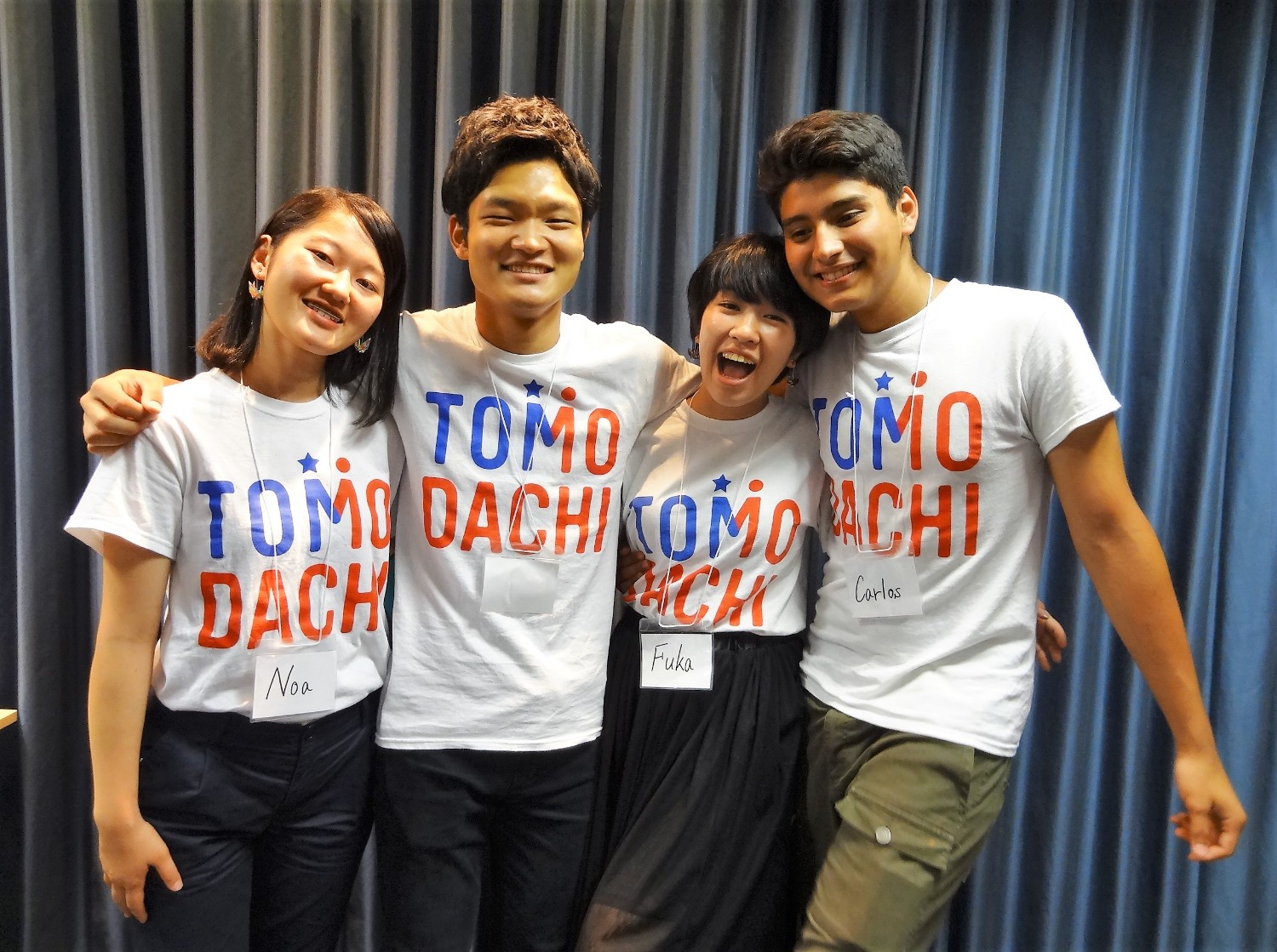 「TOMODACHI」のTシャツを着た4人の若者が、名前の書かれた名札を胸に下げて笑顔で肩を組んでいる写真