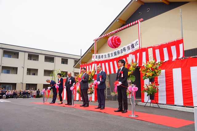 スーツ姿の男性たちが紅白の幕で飾られた建物の前で並び、赤いテープカット用のリボンを持って復興住宅の完成を祝う式典に参加している様子を写した写真