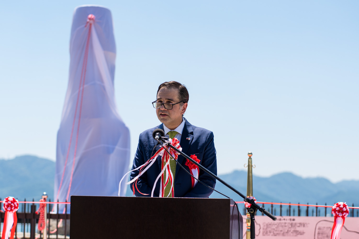 白い布に赤い紐がかけられたモニュメントの前で青いスーツに黄緑のネクタイを着けた男性が演台の前でマイクに向かって話している、晴天の記念式典の写真