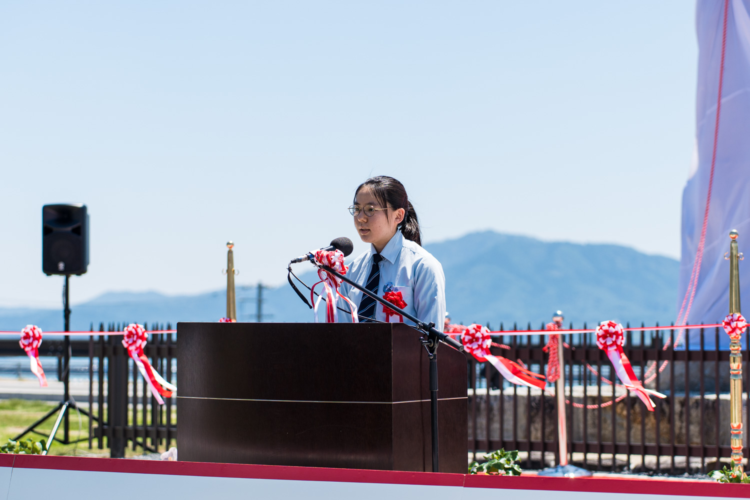 赤白のリボンで飾られた柵や白布に包まれたモニュメントの一部が見える前で、眼鏡をかけた若い女性が水色のシャツにネクタイ姿で演台に立ち、マイクで話している屋外での記念式典の写真
