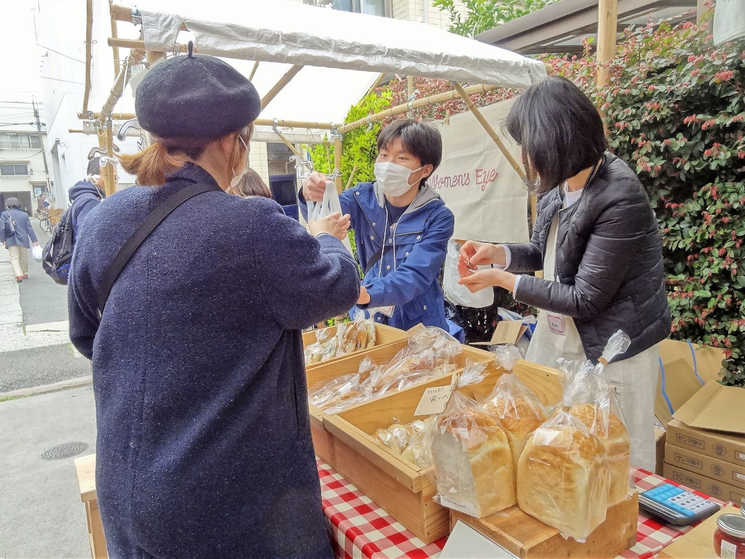 焼き立てのパンが並ぶブースで、マスクをした男性スタッフが商品が入った袋を女性客に渡そうとしており、女性スタッフがお釣りを準備している写真