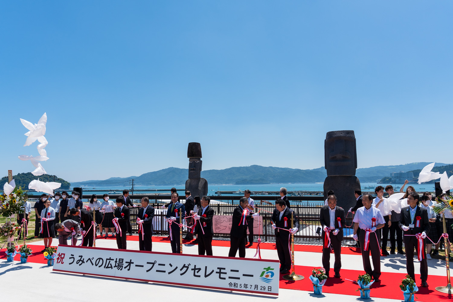 青空の下、海と山を背景に赤いカーペットが敷かれた広場でモアイ像の前に複数の人々が並び、『うみべの広場オープニングセレモニー』の看板の前でテープカットをしている写真