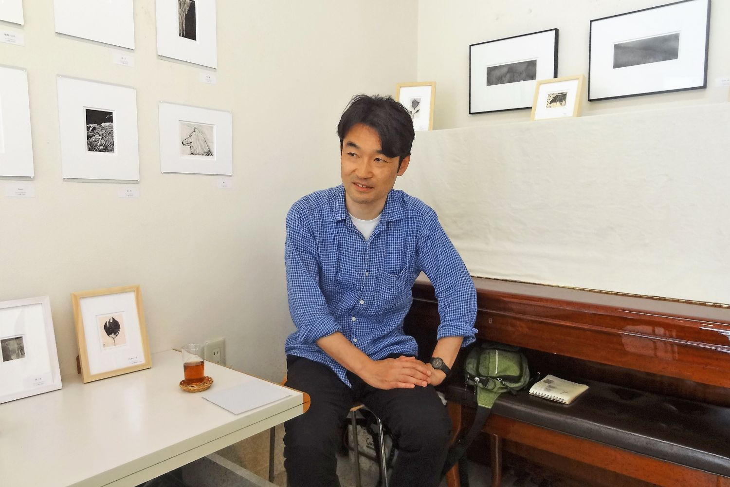テーブルの上や壁に複数の額入り作品が飾られている室内で、椅子に座って話している青いシャツを着た谷村さんの写真