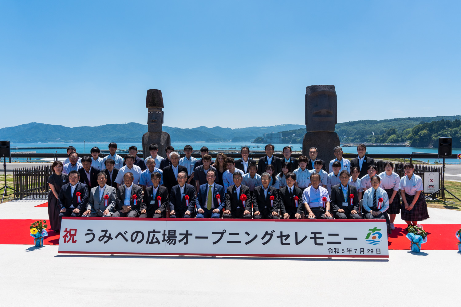 「祝 うみべの広場オープニングセレモニー」と書かれた看板の前でスーツ姿の大人たちと制服姿の学生たち約40人が赤い絨毯の上で並んで記念撮影した写真