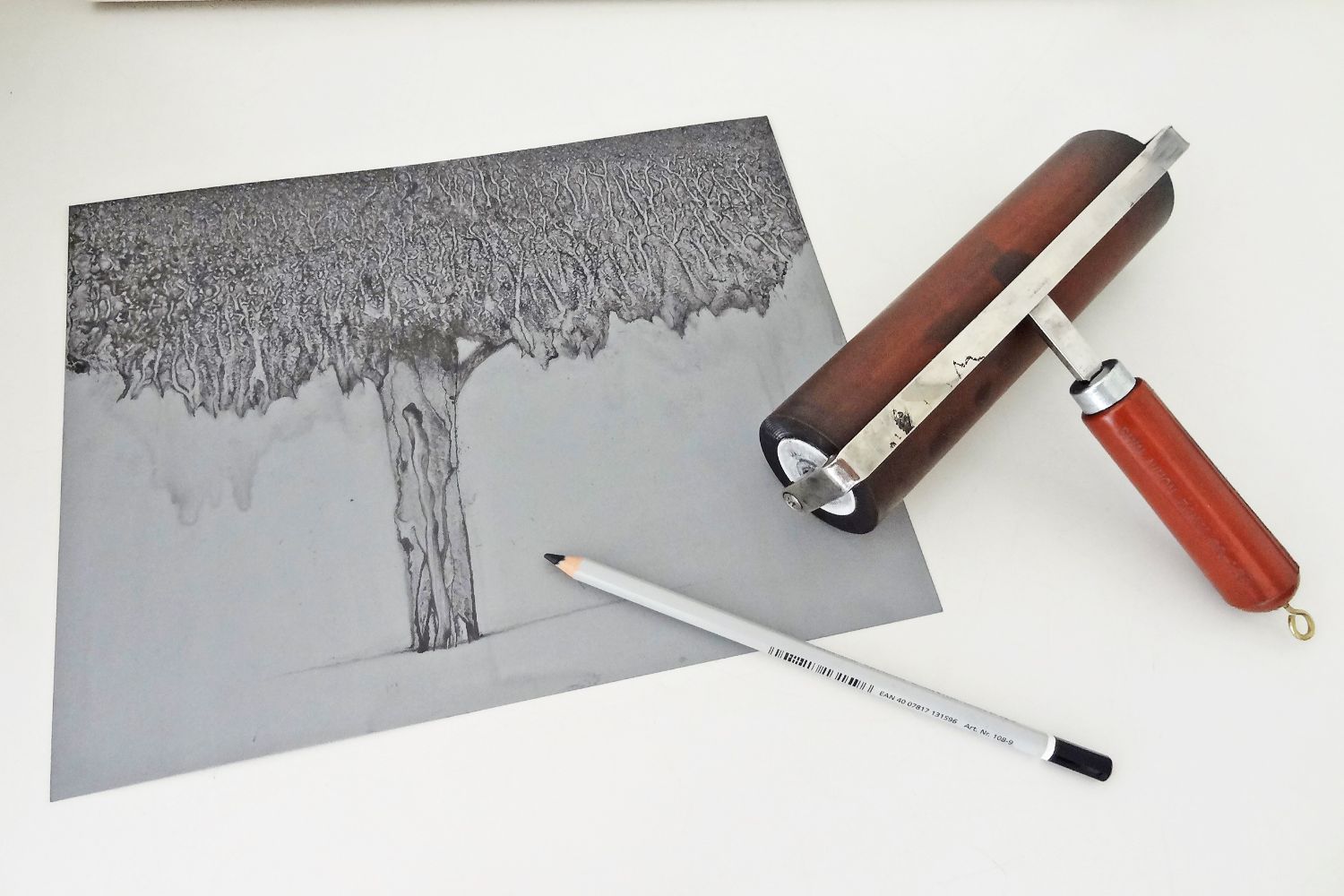 紙の上に鉛筆で描かれた一本の大きな木の絵があり、その隣には鉛筆と、版画制作に使われる木製の持ち手がついたインクローラーが右側に置かれている写真