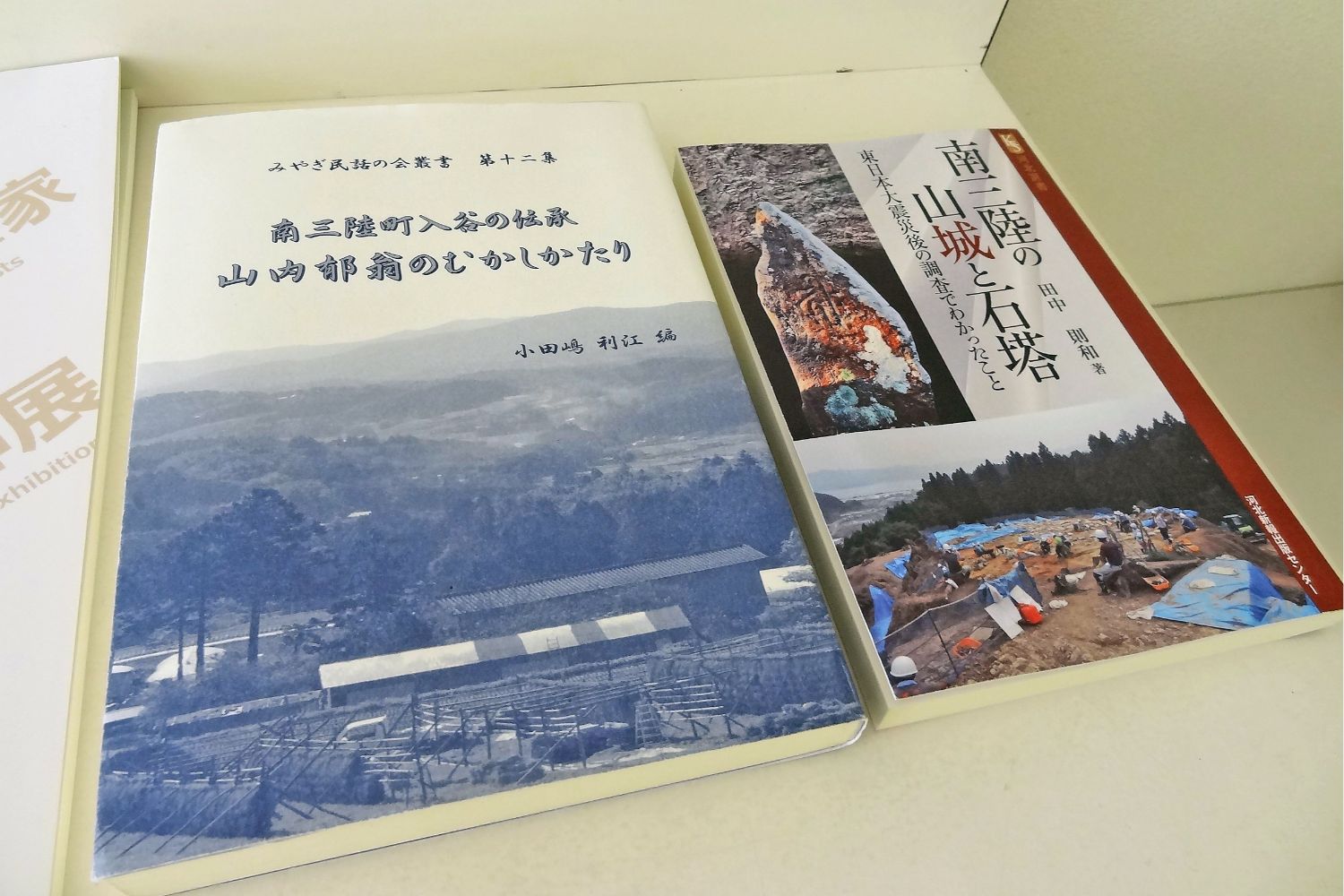 左側の本は青みがかった山や木々の風景写真が表紙に使われ、「南三陸入谷の伝承 山内郁翁のむかしかたり」と書かれており、右側の本は考古学的な発掘現場と遺物の写真が表紙に使われ、「南三陸の山城と石塔」と書かれている写真