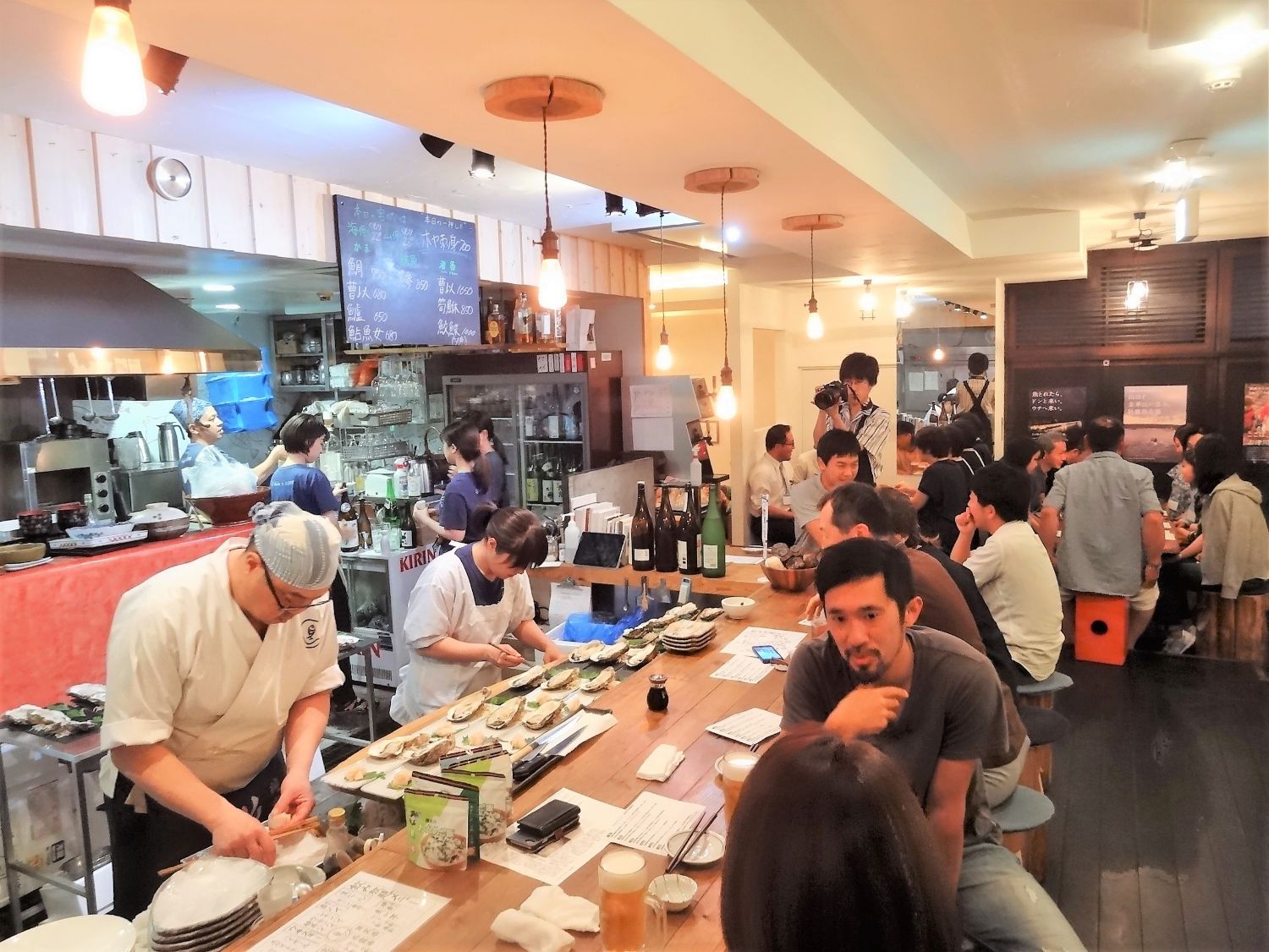 調理場で料理人が真剣に盛り付けをしている一方で、カウンター席には食事を楽しむ人々が座っている写真