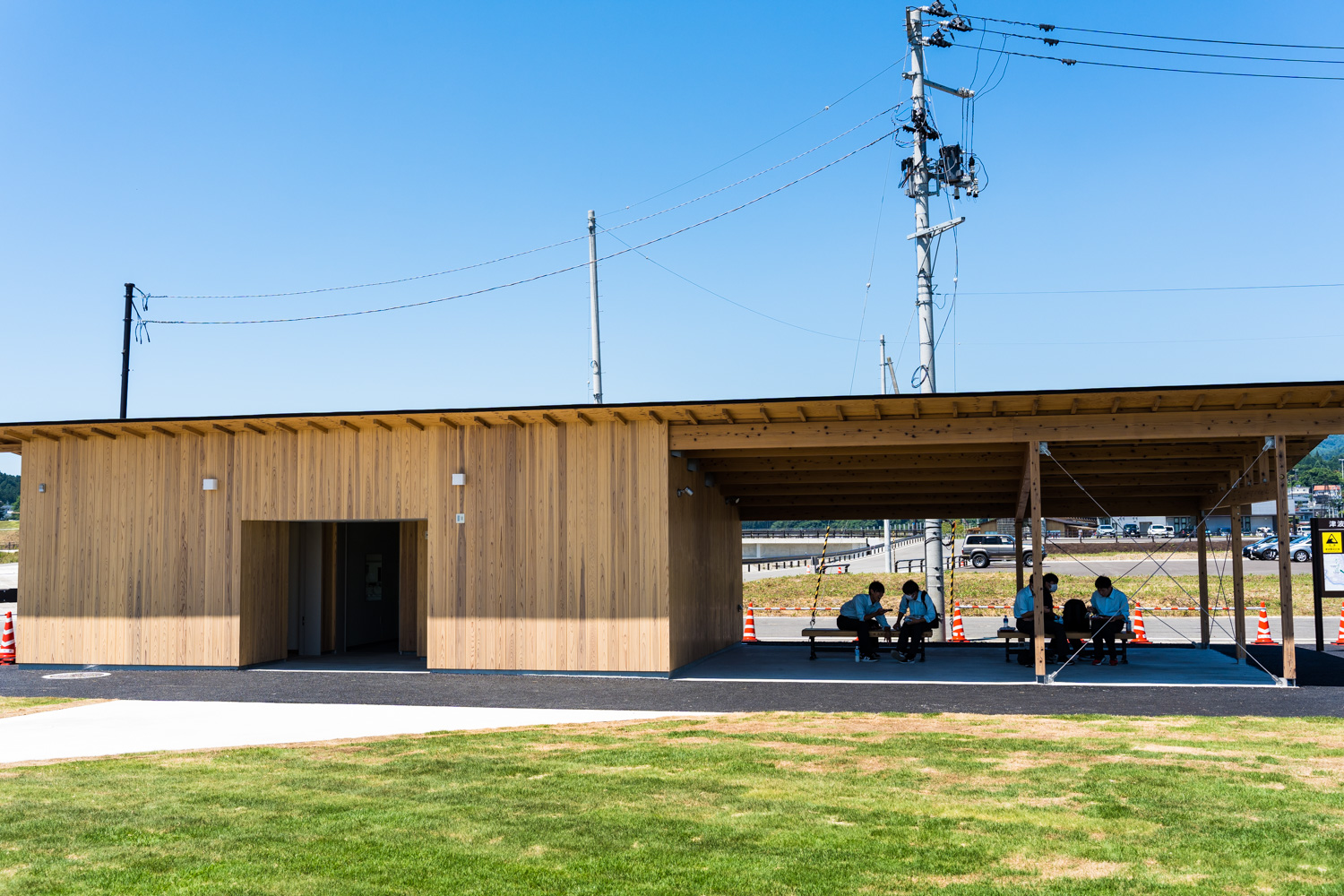 青空の下、芝生の広場に面した木造の建物があり、右側の屋根付きベンチに数人が座っている写真