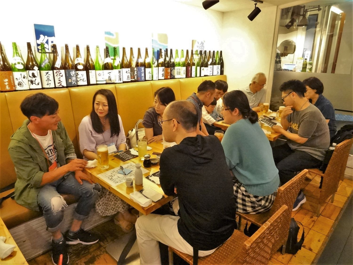 背後には日本酒の瓶がずらりと並んでいる黄色いソファー席に、10人ほどの男女が座り飲み物を片手に談笑している写真
