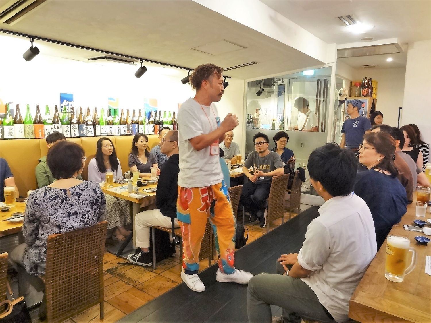 オレンジ色の大漁旗柄のズボンを履いた千葉豪さんが立ち上がって話しており、周囲の客がその話に耳を傾けている写真