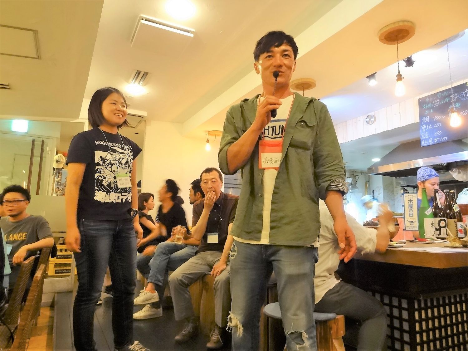 マイクを持った高橋直哉さんとその隣の成影沙紀さんが中央に立ち、周囲の席に座っている参加者に向かって話している様子の写真