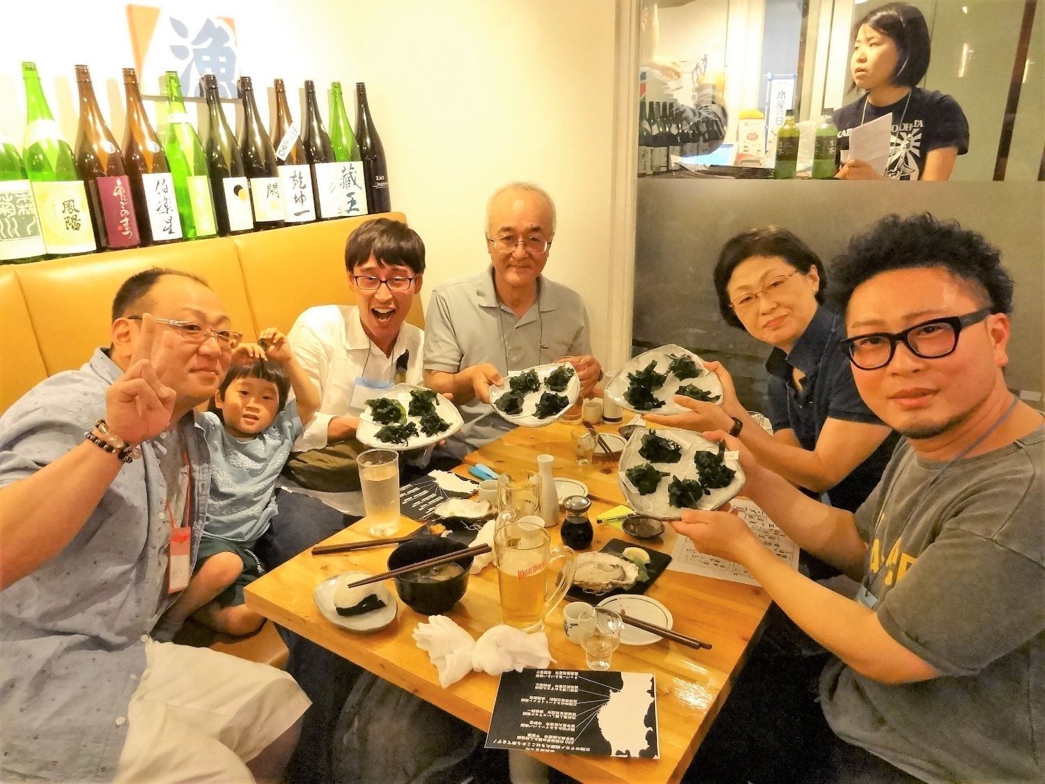 家族連れを含む6人のグループが、ワカメの皿を手に持ってカメラに向かって笑顔でポーズを取っている写真