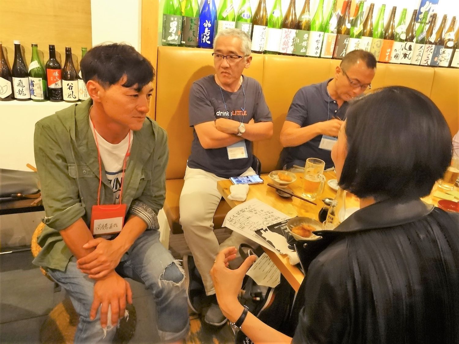 年配の男性たちが食事を楽しむ店内で、黒髪の女性が話しかけるのを、緑の上着と名札をつけた高橋直哉さんが少し緊張した表情で聞いている写真