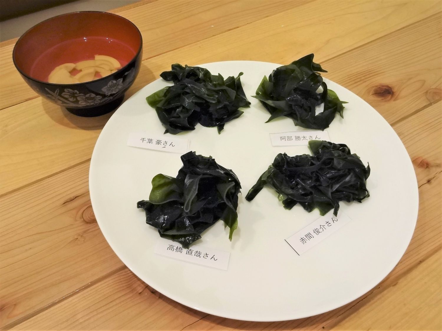 吸い物が入っているお椀と、白い皿に4人の名前が書かれた札の上に、それぞれ異なる見た目のワカメが盛られている写真