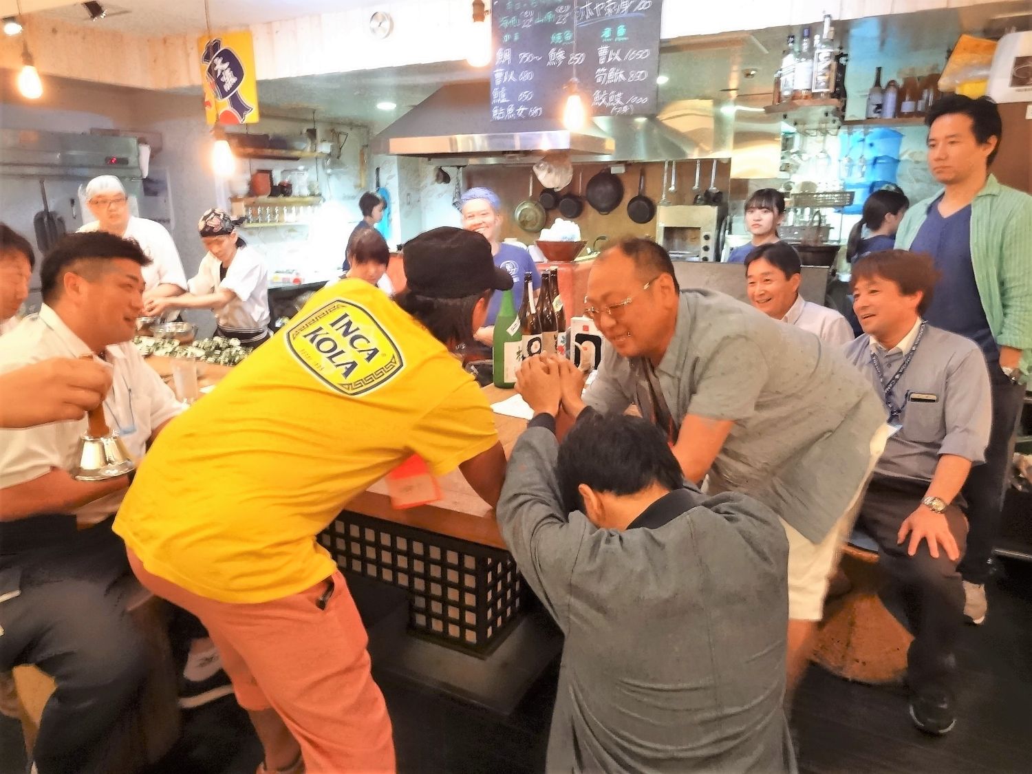居酒屋のカウンター席で2人の男性が真剣な表情で腕相撲を開始しようとしており、周囲の人々が楽しそうに見ている写真