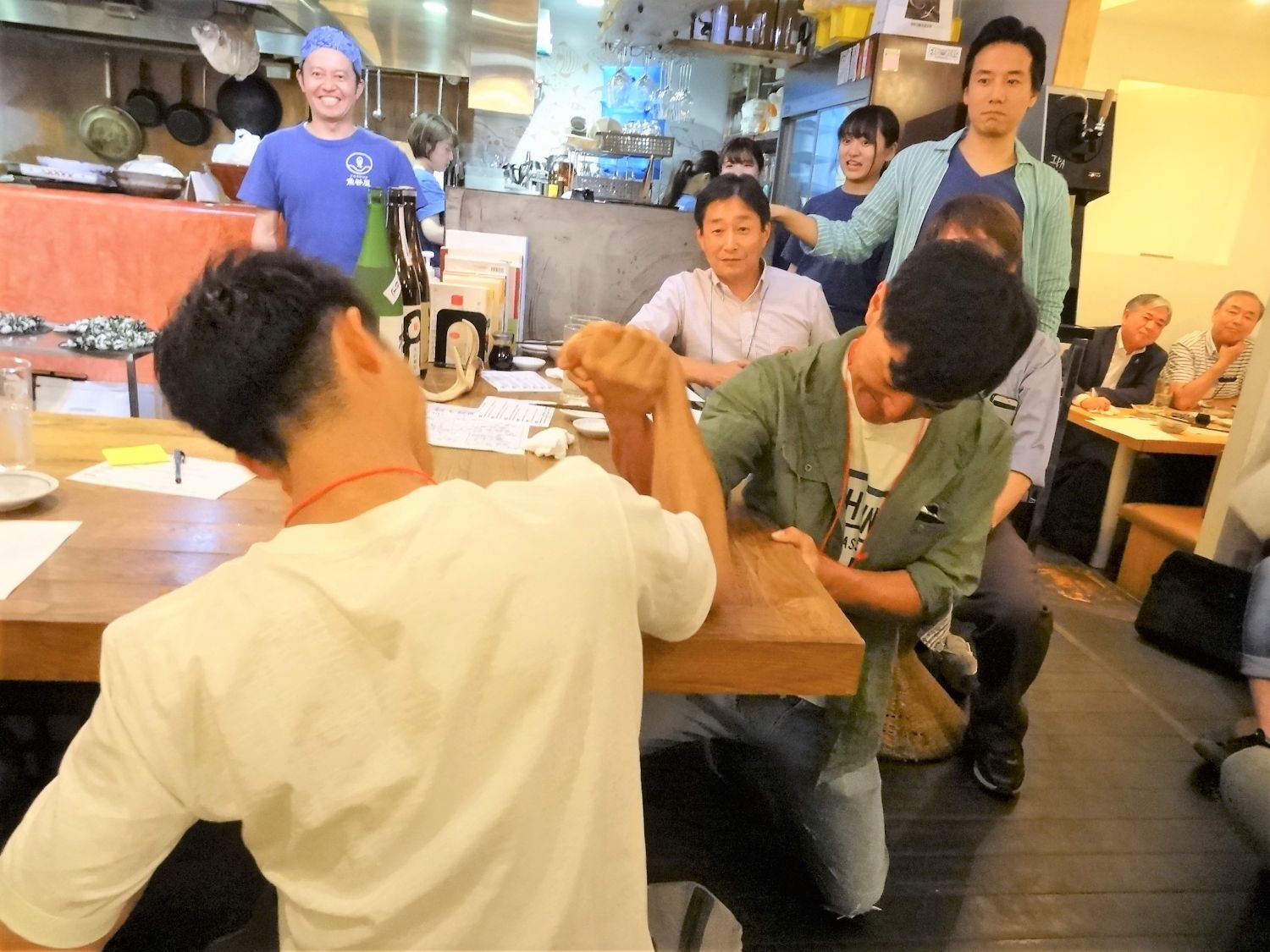 周囲で観客が見ているなか、木製のカウンターで2人の男性が腕相撲をしているところを後ろから撮影している写真