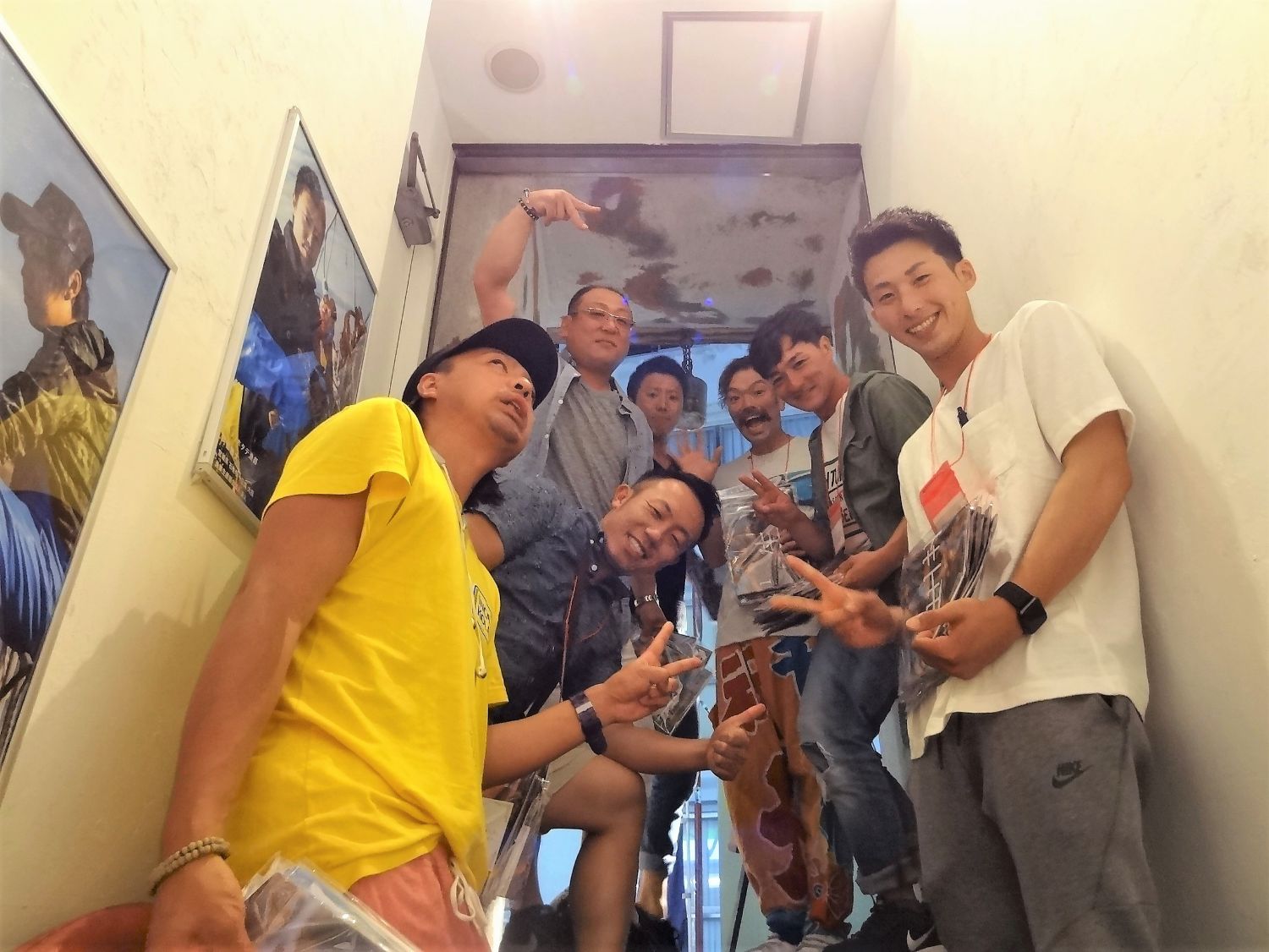 白い壁に囲まれた階段で、8人の男性たちがにこやかにピースサインをしたりポーズを取っている写真