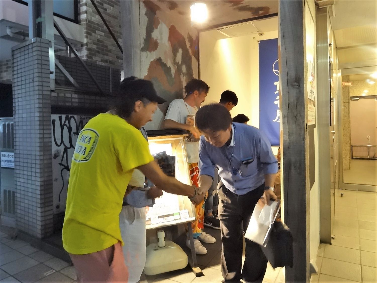 店の入り口前で、黄色いTシャツを着てキャップをかぶった男性が、参加者の男性を握手をして見送っており、奥には他の参加者たちが立っている、交流会終了後の店先の写真