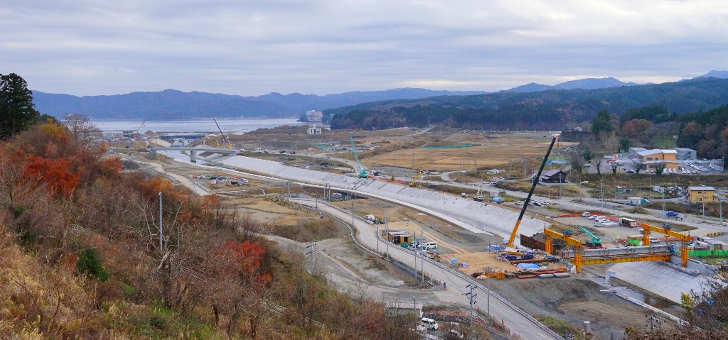 遠くに山々や海が見え、手前の方にはクレーンなどを使った工事が行われている場所が写っている写真