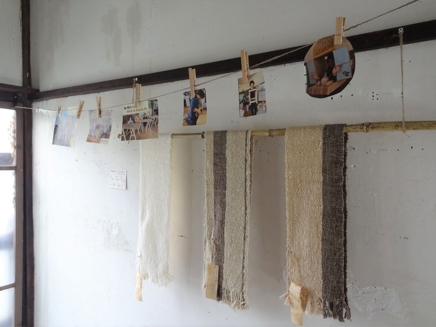 白い壁に沿って上部に6枚の写真が飾られ、写真の下に手造りのマフラーが竹の棒に掛けられ展示された様子の写真