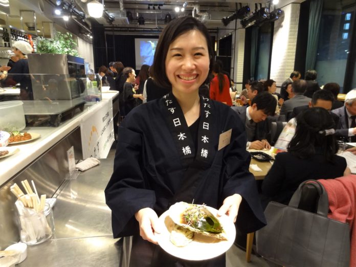 「わたす日本橋」と書かれた紺色の法被姿の女性が笑顔で牡蠣の蒸し焼き料理が盛り付けられたお皿を持っている写真