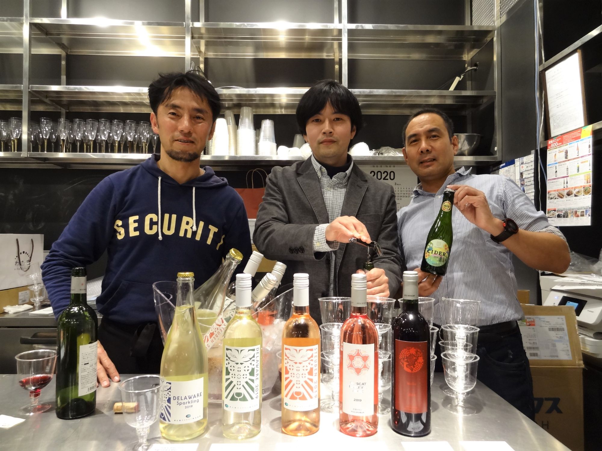 重ねて置かれたワイングラスや5本のワインボトルが並んでいる傍に立つ、ワイナリー代表の3人の写真