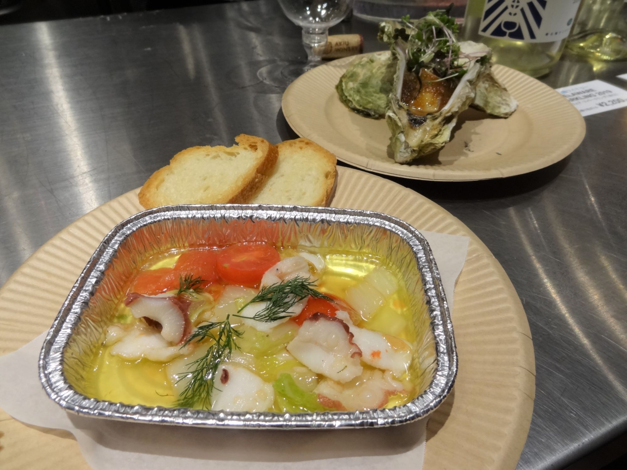 ベージュ色の紙皿の上に2切れのスライスしたフランスパンと完成した「南三陸産 タコとホタテのアヒージョ」、「南三陸産 戸倉っこかきの蒸し焼き」をアップで撮影した写真