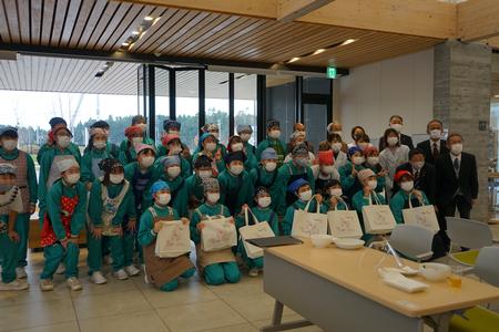 緑色のジャージにエプロンを着て、頭にバンダナを付けた歌津中学校の生徒達と白衣を着た共立女子大学生、関係者の方々が集まり記念撮影をしている写真