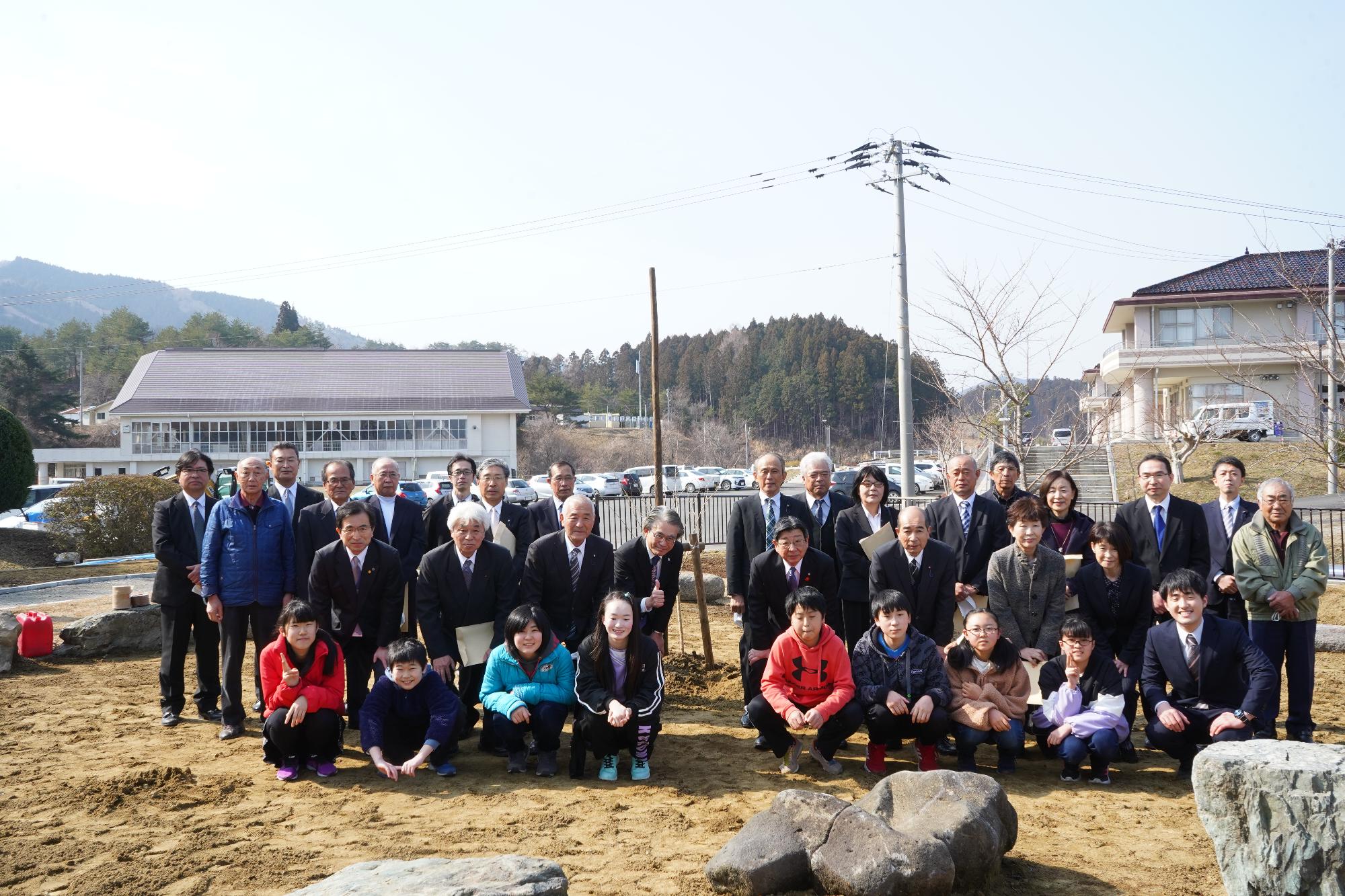 スーツ姿の大人たちとカラフルな服を着た子どもたちが、新しく植えられた若木の前で記念撮影をしている写真