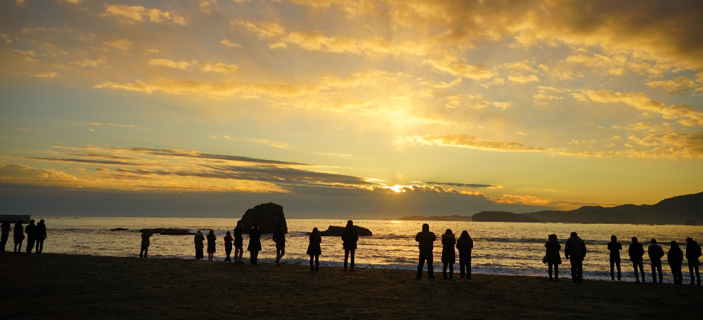 金色に輝く空と海を海岸から眺めている沢山の人のシルエットが写っている写真