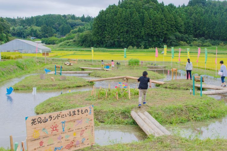 田んぼの中に作られた木の橋を子どもたちが渡って遊んでおり、周囲には自然観察用の看板や旗が立ち、奥には棚田と森林が広がる風景の中で里山体験が行われている様子の写真