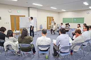 ホワイトボードの前に立つ男性が説明をしていて、それを囲むように十数人の男女が円形に椅子に座って資料を持ち、真剣に話を聞いている様子を写したグループワークの写真
