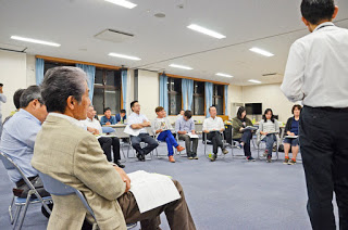 窓のある明るい会議室で、十数人の男女が円形に座って話し合いをしており、手前に座る年配の男性の後ろ姿が写っている、講師らしき男性が立って話している様子を写した話し合いの写真