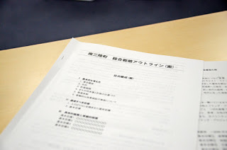机の上に置かれた「防災対策 総合検討アウトライン（案）」という表題の日本語の資料がクローズアップされており、災害対策に関する項目が並んでいる様子を写した資料の写真