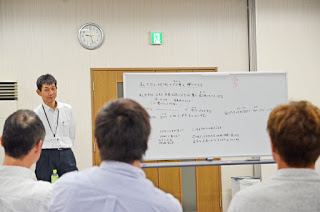 壁掛け時計と非常口のドアの前にホワイトボードが置かれ、そこに書かれた内容を前にした男性講師が立って説明し、数名の男性が背を向けて真剣に聞いている様子を写した説明中の写真