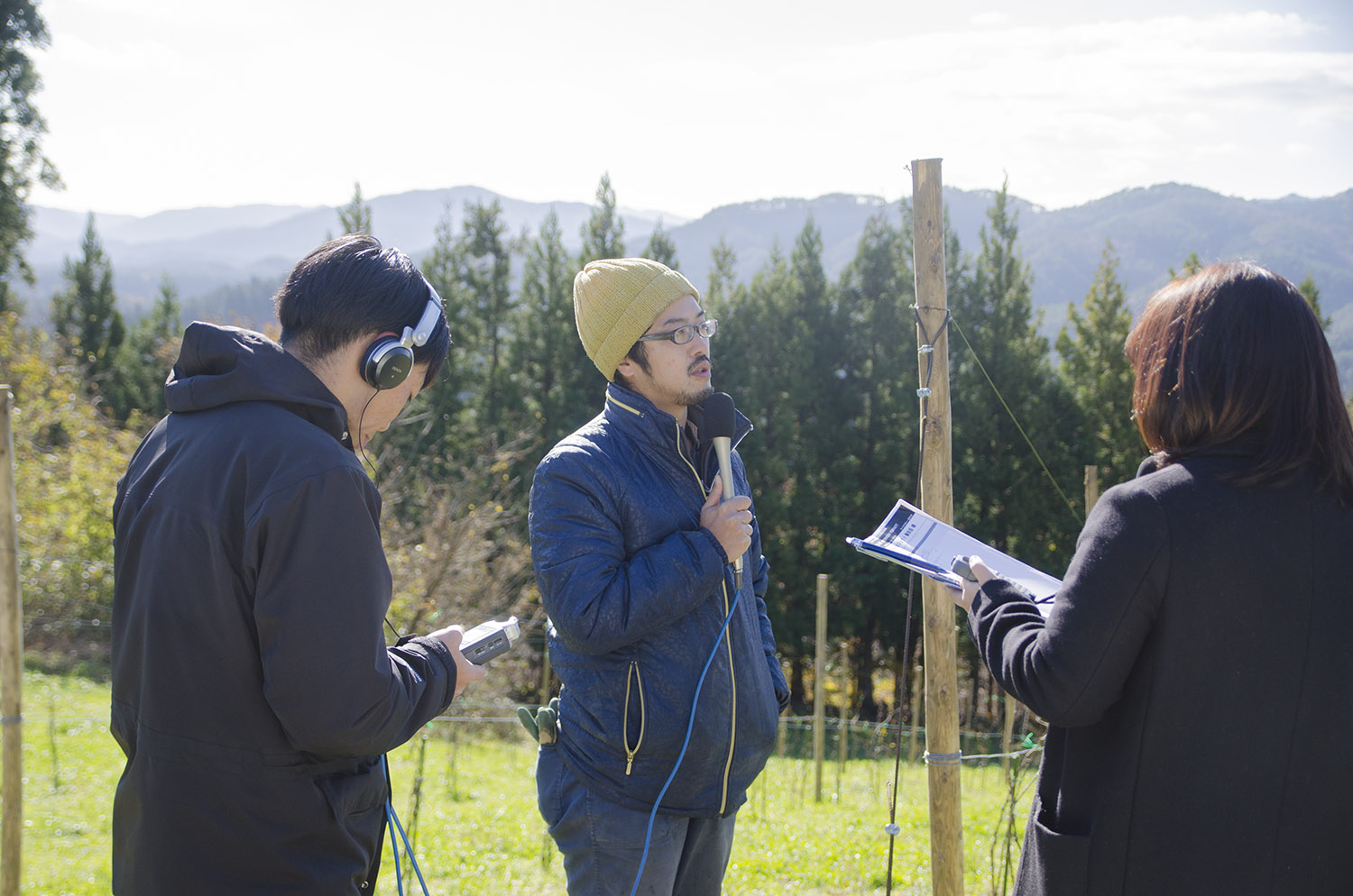 山と森を背景にした農園で、機材を持った男性とリポーターの女性と向かい合って立ち、マイクを持った藤田さんがインタビューを受けている写真
