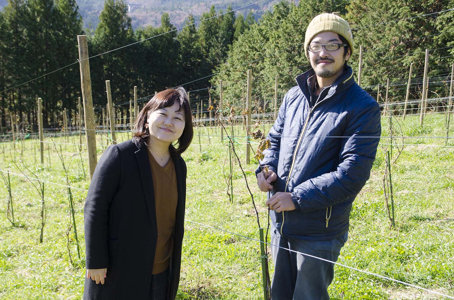 ブドウ畑で、ブドウの枝を持ちながら微笑んでいる藤田さんと、女性リポーターが並んで立っている写真