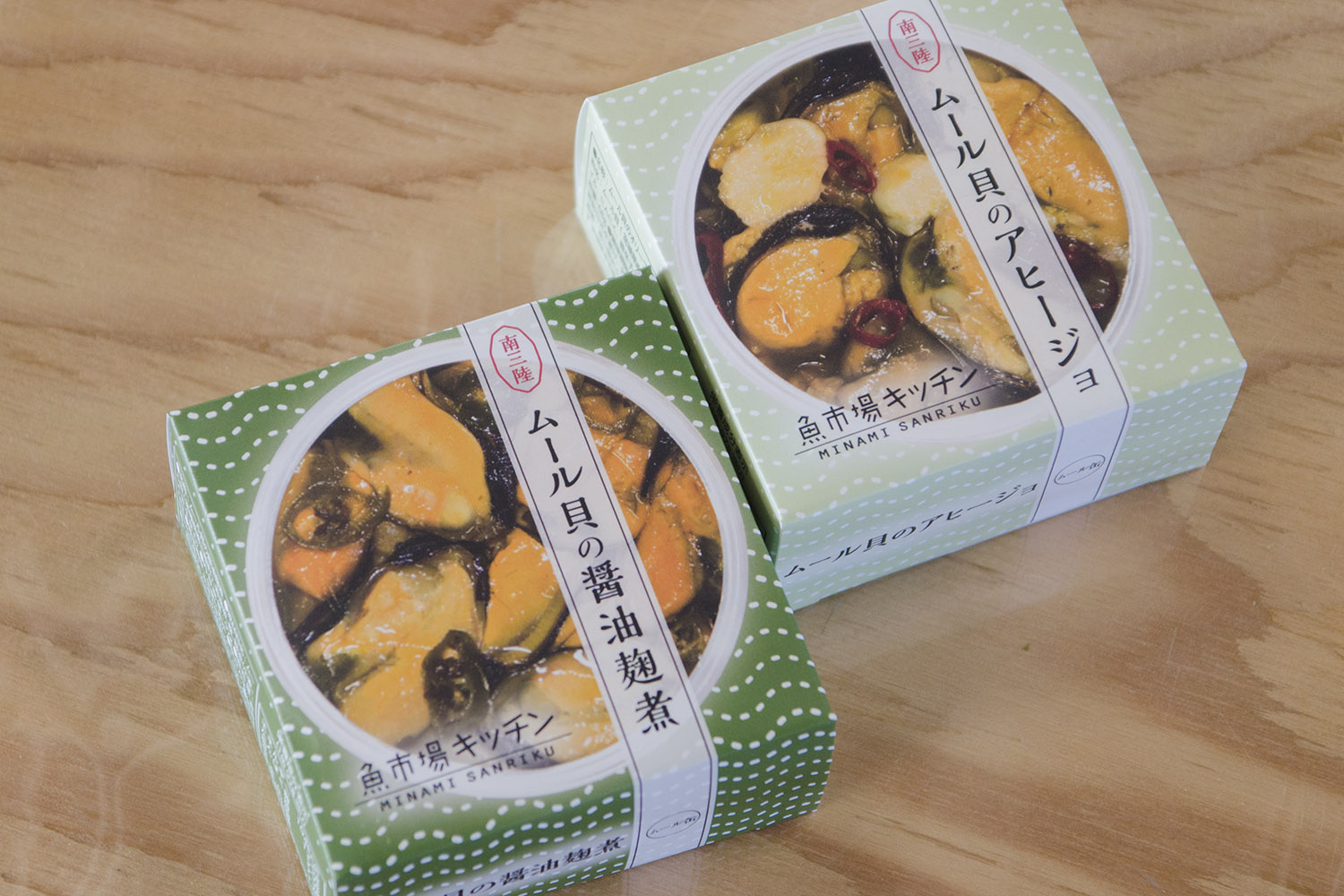 木のテーブルの上にムール貝の料理の写真が大きく印刷され「ムール貝の醤油麹煮」と「ムール貝のアヒージョ」と書かれた緑色の箱が2つ並べられている写真