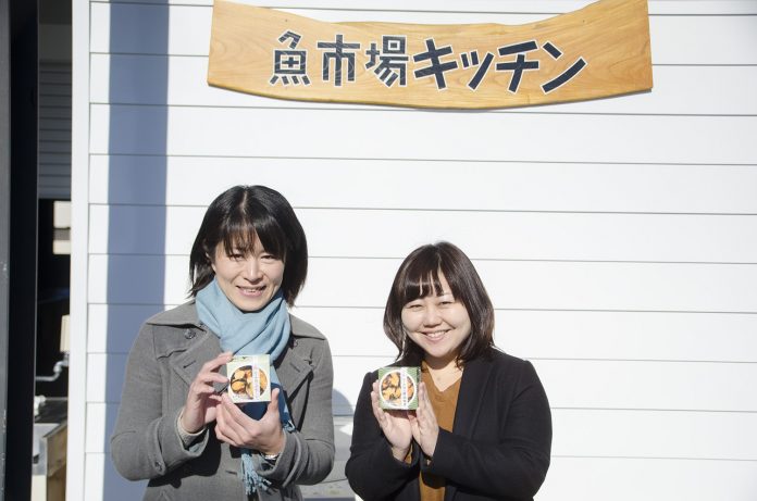 「魚市場キッチン」と書かれた木の看板の前で、グレーのコートを着た中村さんと女性リポーターが並んで立ち、それぞれ手に持ったムール貝料理の缶詰を見せている写真