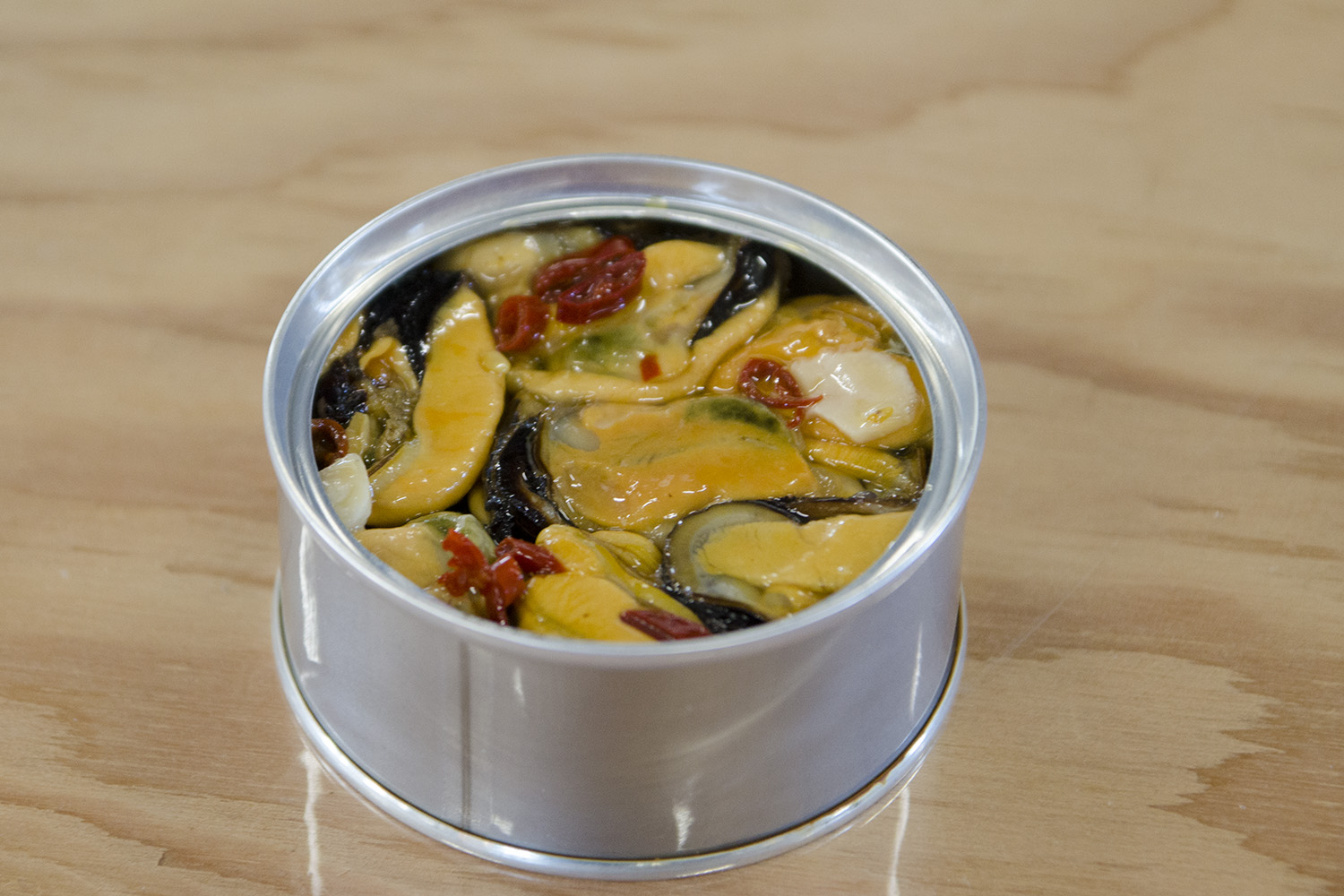 テーブルの上にフタを開けたムール貝の缶詰が置かれており、中にはオレンジ色のムール貝や赤い唐辛子などの具材がぎっしり詰まっている写真