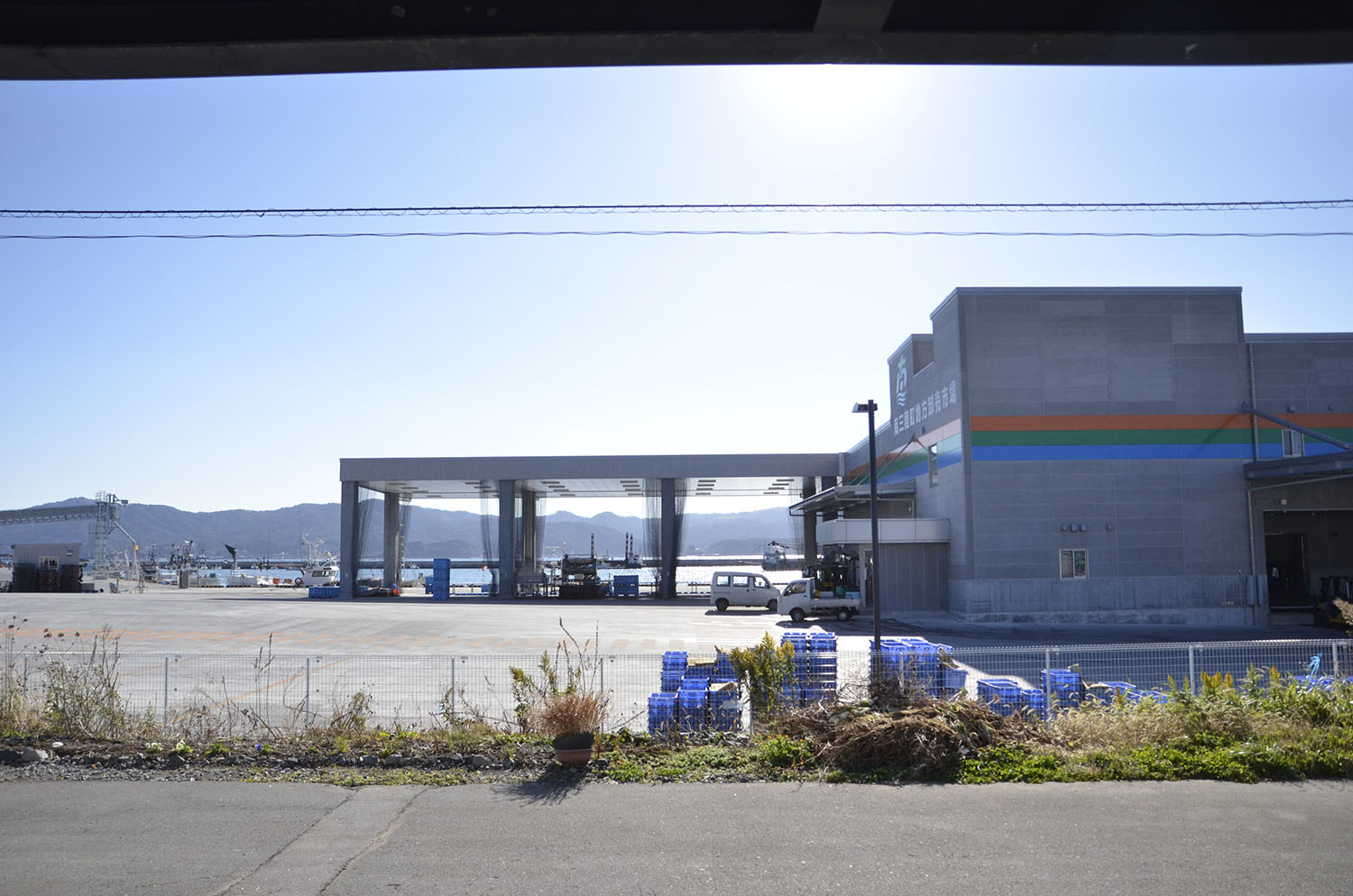 青空の下、漁船が停泊している港があり、海辺にある市場の前に大きな加工場の建物が建っている写真