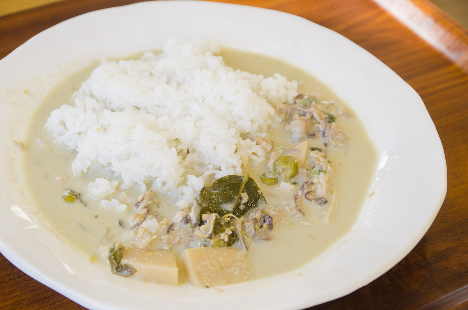 白い皿に盛り付けられた三陸産の牡蠣と野菜が入ったグリーンカレーと白ご飯が、木製のトレイの上に置かれている写真