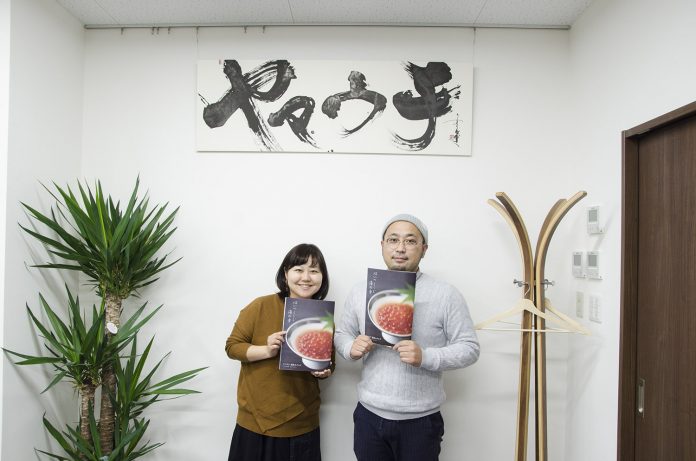 壁には筆文字で書かれた『ヤマウチ』の看板が掛かっている室内で、女性リポーターと灰色のセーターを着た山内さんが、いくらの写真が表紙のカタログを手に、並んで写っている写真