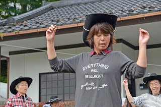 「EVERYTHING WILL BE ALL RIGHT」と書かれた服を着た女性がカウボーイハットをかぶり、他の人たちと一緒に踊っている写真