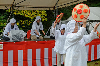 白い頭巾と衣装を着た人々が赤い飾りのついた丸い道具を手に踊っており、後ろの舞台では演奏者が和太鼓をたたいている写真