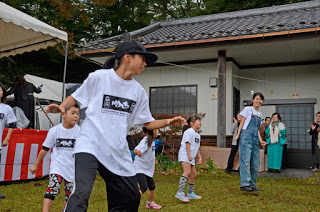 白いTシャツと黒い帽子をかぶった子どもたちが屋外のステージ前で元気にダンスを踊っており、後ろには見物している人々がいる写真